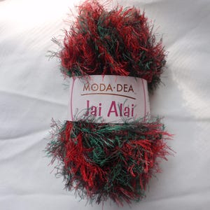 Pode incluir: Uma bola de fio Moda Dea Jai Alai em vermelho, verde e preto. O fio tem uma aparência difusa e texturizada. O rótulo no fio diz "Moda Dea Jai Alai". O fio é provavelmente usado para projetos de tricô ou crochê.