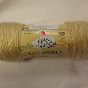 May include: A skein of Yarn Bee Soft Secret yarn in a light yellow color. The label features the brand name, floral accents, and the text "Soft Secret". The yarn is tightly wound and ready for knitting or crocheting projects.