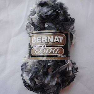 May include: A skein of Bernat Boa Nouveau yarn in shades of gray, black, and white. The yarn has a fluffy, textured appearance, with the brand name and product name printed on a label wrapped around the center.