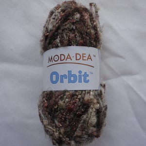 Pode incluir: Um novelo de fio Moda Dea Orbit em tons de castanho, creme e vermelho com detalhes metálicos. O fio é embrulhado com uma etiqueta branca que diz "MODA-DEA" e "Orbit" em texto azul.
