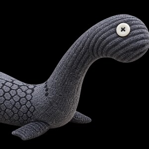 May include: A plush toy shaped like a long-necked aquatic creature, crafted from dark gray ribbed fabric. It features a white button eye with four holes, and a dark gray geometric scale pattern printed along its back and sides.