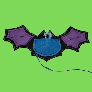 May include: A felt bat-shaped needle holder in black, purple, and blue. The wings are purple with black trim, and the center has a blue pocket with needles and thread. A blue ribbon extends from the pocket. The background is green.