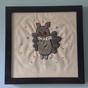 Op de afbeelding: Een vierkante zwarte lijst met een handgeborduurd werk op gebroken wit doek. Het ontwerp toont een grijs, gevleugeld wezen met rode ogen en een witte ruchekraag, omringd door kleine, verspreide, veelkleurige geborduurde stippen.