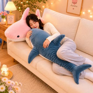 May include: A blue shark-shaped cushion is being held. A large, pink shark cushion rests behind it. The cushions are on a beige sofa. The shark cushions have fins, tails, and a friendly expression.