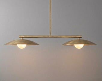 May include: A linear pendant light fixture featuring a horizontal brass bar suspended by a single vertical rod. Two wide, shallow, saucer-shaped brass shades are attached to the ends, each housing a small, glowing, spherical white glass bulb.