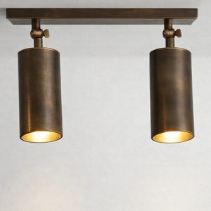 May include: A dual-head ceiling spotlight fixture featuring a rectangular mounting plate and two cylindrical heads in a dark bronze finish. Each head is attached to the base with an adjustable swivel joint, emitting a warm light from the bottom.