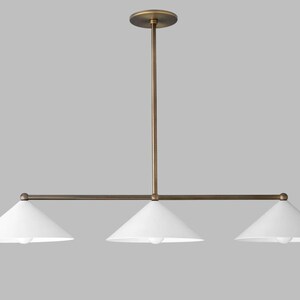 A linear chandelier featuring a central brass rod and a matching horizontal bar that supports three white conical shades. The fixture is suspended by a single vertical brass stem attached to a circular brass ceiling canopy at the top.