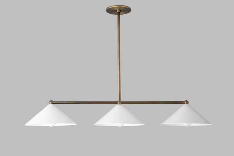 A linear chandelier featuring a central brass rod and a matching horizontal bar that supports three white conical shades. The fixture is suspended by a single vertical brass stem attached to a circular brass ceiling canopy at the top.