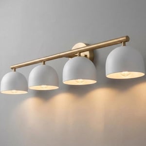 May include: A wall-mounted light fixture featuring a horizontal brass-toned metal bar supporting four matte white dome-shaped shades. Each shade houses a light bulb, casting a warm glow downward against the wall surface behind the fixture.