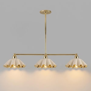 A gold-toned metal chandelier featuring a central vertical rod connected to a horizontal bar that supports three downward-facing, cream-coloured, scalloped-edge shades. Each shade has a gold-toned rim and an exposed light bulb inside.