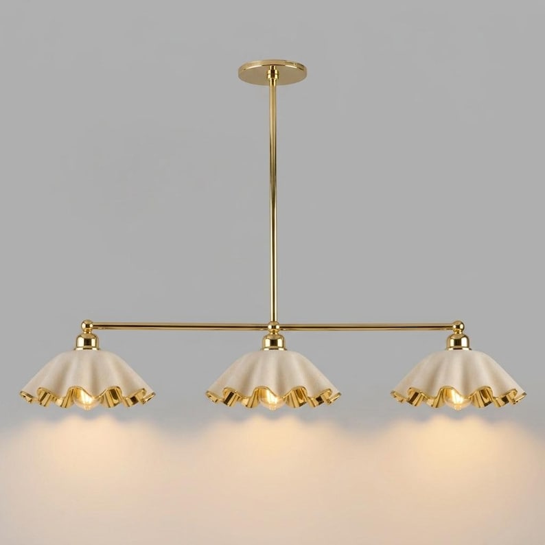 A gold-toned metal chandelier featuring a central vertical rod connected to a horizontal bar that supports three downward-facing, cream-coloured, scalloped-edge shades. Each shade has a gold-toned rim and an exposed light bulb inside.