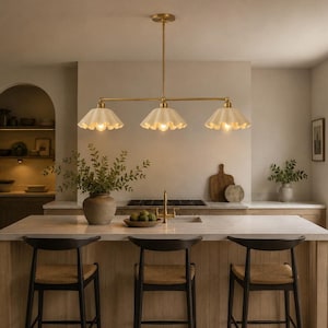 May include: A linear pendant light fixture featuring a long brass rod suspended from the ceiling, holding three downward-facing, scalloped, off-white glass shades. Each shade contains a single light bulb, providing warm illumination over the kitchen island.