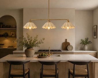 May include: A linear pendant light fixture featuring a long brass rod suspended from the ceiling, holding three downward-facing, scalloped, off-white glass shades. Each shade contains a single light bulb, providing warm illumination over the kitchen island.