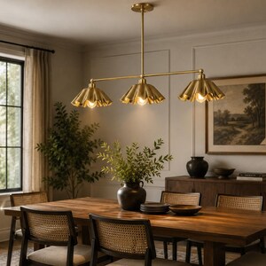 A linear chandelier featuring three brass-toned metal shades with a pleated, scalloped edge design. The fixture is suspended by a central vertical rod and a horizontal bar, with each shade housing a single light bulb facing downward.