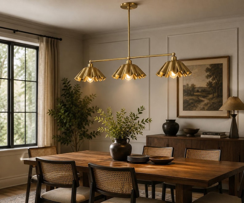 A linear chandelier featuring three brass-toned metal shades with a pleated, scalloped edge design. The fixture is suspended by a central vertical rod and a horizontal bar, with each shade housing a single light bulb facing downward.