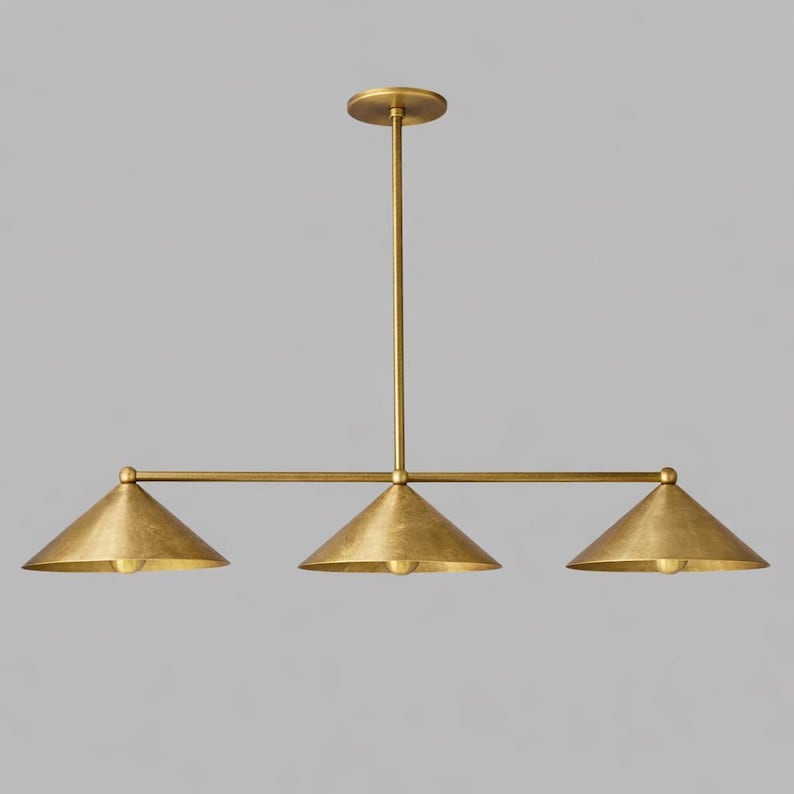 A linear chandelier featuring a central vertical rod connected to a horizontal bar that supports three downward-facing conical shades. The entire fixture is finished in a brushed brass tone with a smooth, metallic surface texture.