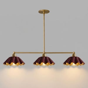 A linear chandelier featuring a central brass rod and three downward-facing, ruffled-edge shades in a deep burgundy finish with gold interiors. Each shade houses a single light bulb, casting a warm glow from the fixture.