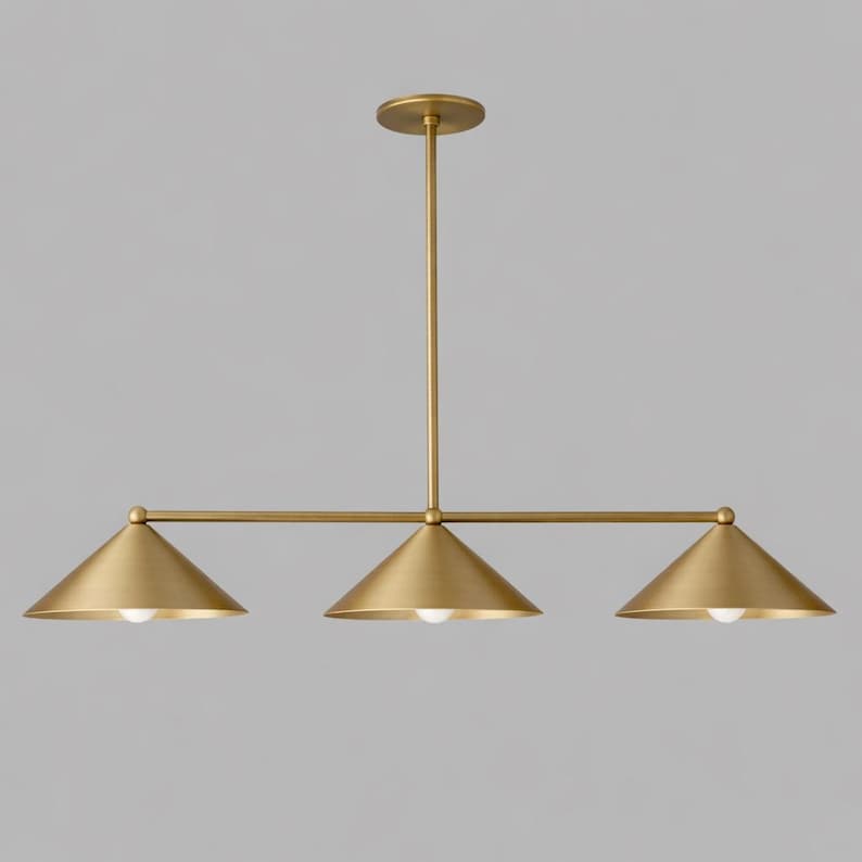 A linear pendant light fitting with a brushed brass finish, featuring a central vertical rod connected to a horizontal bar that supports three downward-facing conical shades, each containing a single small white light bulb.