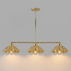 A gold-toned metal chandelier featuring a central vertical rod connected to a horizontal bar. Three scalloped, bell-shaped shades are attached to the bar, each housing a light bulb that emits a warm, soft glow downwards.