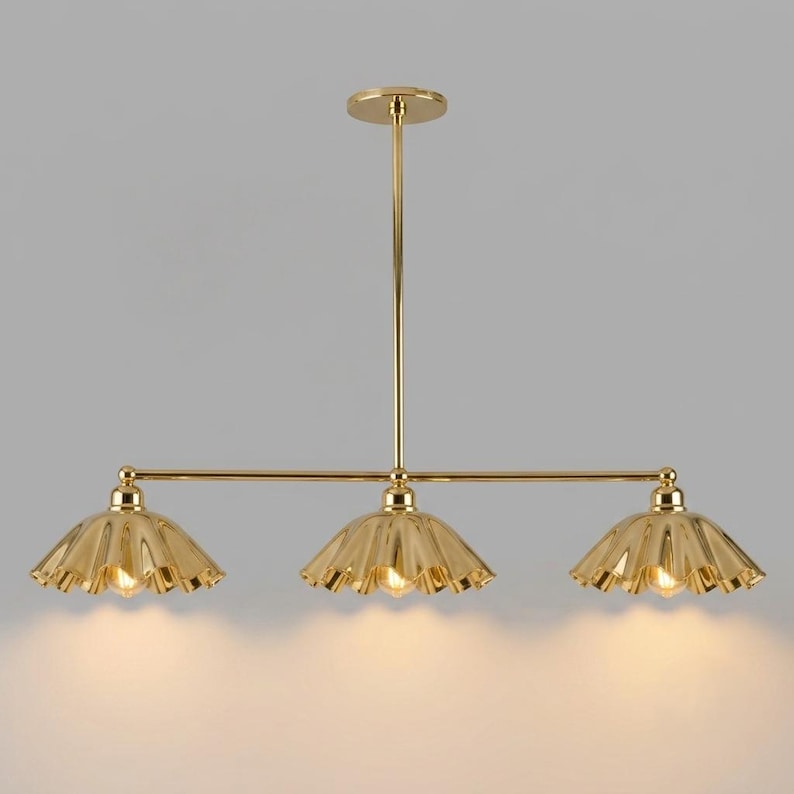 A gold-toned metal chandelier featuring a central vertical rod connected to a horizontal bar. Three scalloped, bell-shaped shades are attached to the bar, each housing a light bulb that emits a warm, soft glow downwards.