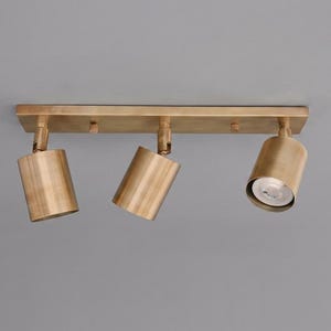 Puede incluir: Una lámpara de techo con tres focos y acabado en latón cepillado. La placa de montaje rectangular sostiene tres cabezales cilíndricos ajustables, cada uno con un exterior de tono dorado metálico y una abertura circular para una bombilla.