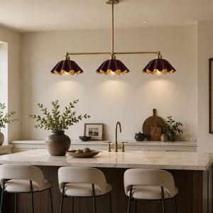 A linear chandelier featuring three dark burgundy, pleated, scalloped-edge shades with gold-toned interiors, suspended from a central brass-finished rod. The fixture provides downward lighting over a kitchen island.