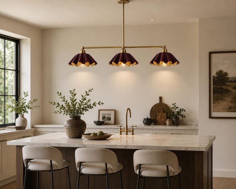 A linear chandelier featuring three dark burgundy, pleated, scalloped-edge shades with gold-toned interiors, suspended from a central brass-finished rod. The fixture provides downward lighting over a kitchen island.