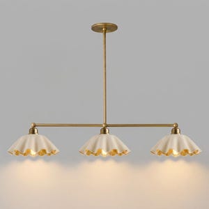 A linear chandelier featuring a central brass rod and three downward-facing, scalloped-edge off-white shades. The fixture is suspended by a single vertical brass stem connected to a circular ceiling canopy, emitting a warm light.