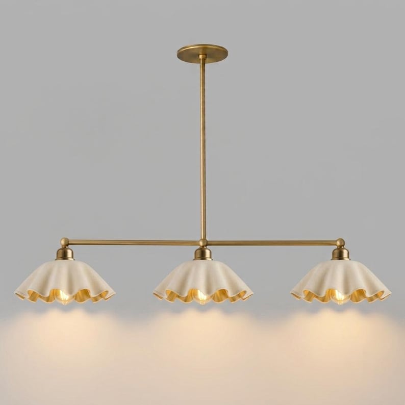 A linear chandelier featuring a central brass rod and three downward-facing, scalloped-edge off-white shades. The fixture is suspended by a single vertical brass stem connected to a circular ceiling canopy, emitting a warm light.