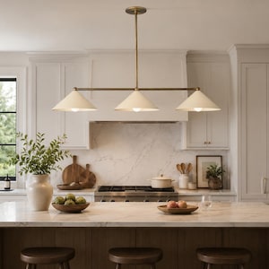 Pode incluir: Uma luminária pendente linear com três cúpulas cónicas em tom branco sujo, suspensas por uma barra horizontal em latão e uma haste vertical fixada a uma base circular no teto, proporcionando iluminação direcionada para baixo.