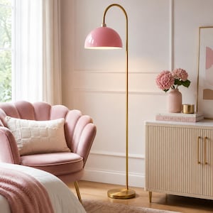 Op de afbeelding: Een vloerlamp met een hoge, slanke goudkleurige metalen standaard en een gebogen hals die een glanzende, koepelvormige roze kap ondersteunt. De lamp heeft een ronde voet en verspreidt een zacht, warm licht van onder de kap naar de omgeving.