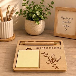 Puede incluir: Un organizador de escritorio de madera con ranura para bolígrafo, espacio cuadrado para notas adhesivas amarillas y un diseño de mariposa grabado con el texto "Que no se me olvide". Incluye un bolígrafo de madera, una taza de cerámica y un marco.
