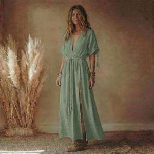 May include: A sage green maxi dress with a deep V-neckline and short sleeves. The dress features a high slit, a rope belt, and a flowing silhouette. The model is wearing brown boots and bracelets.
