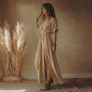 May include: A beige maxi dress with a high slit and short sleeves. The dress has a cinched waist and a flowing skirt. The model is wearing brown boots and bracelets. The background features tall, dried plants.