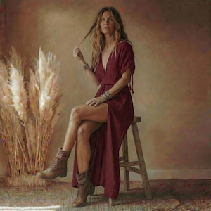 May include: A burgundy maxi dress with a deep V-neck and a high slit. The dress is paired with brown leather boots and multiple bracelets. The model is sitting on a wooden stool, with a backdrop of dried ornamental grass.
