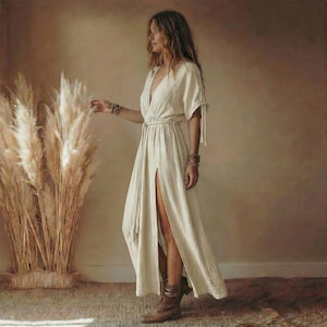 May include: A cream-colored maxi dress with a deep V-neck and a high slit. The dress features short sleeves with tie details and a rope belt. The model is wearing brown boots and several bracelets. Tall, dried pampas grass is in the background.