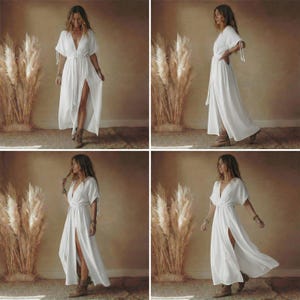 May include: A white maxi dress with a V-neckline and short sleeves. The dress features a high slit on the left side and a tie belt at the waist. The model is wearing brown boots. The dress is made of a lightweight fabric.