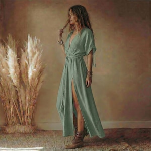May include: A sage green maxi dress with a deep V-neckline and short sleeves. The dress features a high slit on one side and a matching fabric belt. The model is wearing brown boots and bracelets. The dress is made of a lightweight, textured fabric.