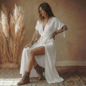 May include: A white, flowing dress with a deep V-neck and short sleeves. The dress features a high slit and is paired with brown boots. The model is seated, with a neutral background and tall, dried plants.