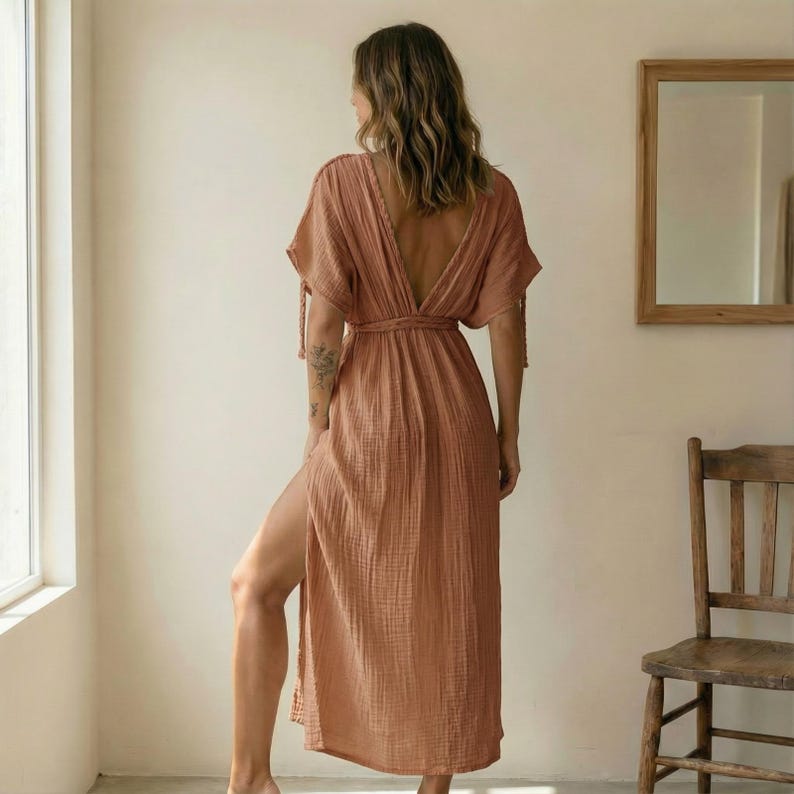 May include: A peach-colored maxi dress with short sleeves and a deep V-neck back. The dress features a waist tie and a high slit on one side, made of a textured fabric. The model is standing near a window.