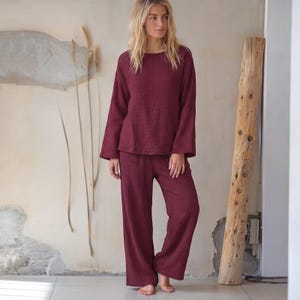 Könnte beinhalten: Ein zweiteiliges Loungewear-Set in tiefem Burgunderrot, bestehend aus einem langärmeligen Oberteil mit Rundhalsausschnitt und einer passenden Hose mit weitem Bein. Der Stoff weist durchgehend eine vertikale Knitterstruktur auf.