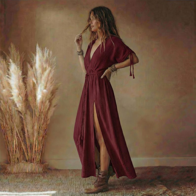 May include: A burgundy maxi dress with a deep V-neck and a high slit. The dress features short sleeves with tie details and a braided belt. The model is wearing brown boots and bracelets. The background includes tall, dried ornamental grass.