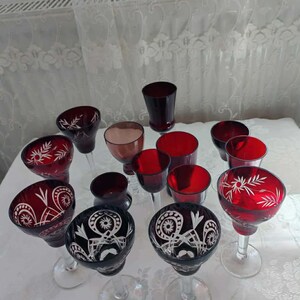 Puede incluir: Un surtido de copas de vino rojas y transparentes. Algunas copas tienen diseños grabados, mientras que otras son de color rojo liso. Las copas varían en forma y tamaño, incluyendo copas de vino y vasos más pequeños. La cristalería está dispuesta sobre una superficie blanca.