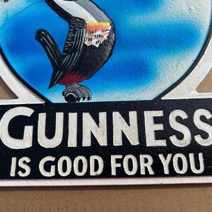 Puede incluir: Un letrero decorativo de metal con una ilustraci&oacute;n colorida de un tuc&aacute;n sobre un fondo azul. Debajo del ave, un panel rectangular negro muestra el texto en relieve blanco GUINNESS IS GOOD FOR YOU. El letrero tiene un borde blanco.