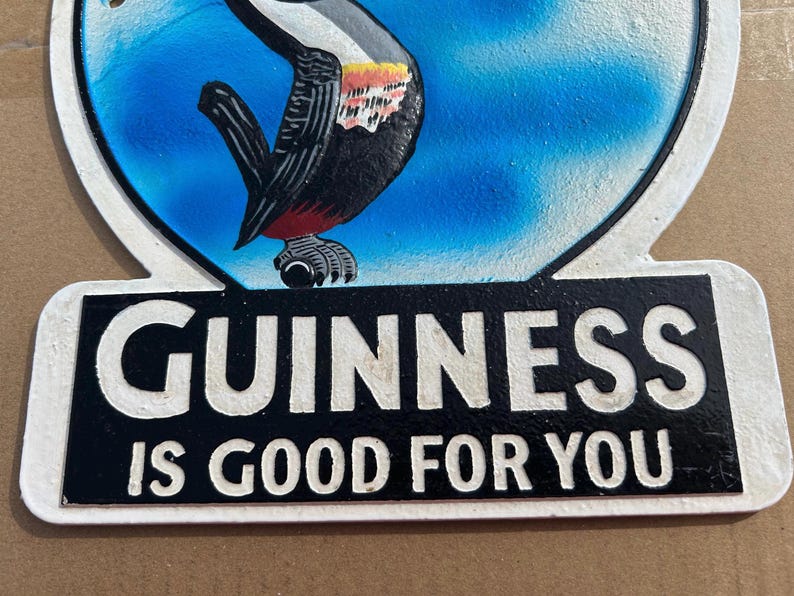 Puede incluir: Un letrero decorativo de metal con una ilustraci&oacute;n colorida de un tuc&aacute;n sobre un fondo azul. Debajo del ave, un panel rectangular negro muestra el texto en relieve blanco GUINNESS IS GOOD FOR YOU. El letrero tiene un borde blanco.
