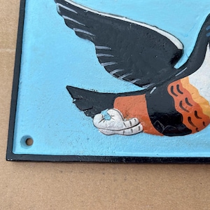 May include: A rectangular metal wall plaque with a light blue background featuring a relief illustration of a toucan. The bird has black wings, a white chest, and orange accents. A small circular hole is visible in the bottom left corner.