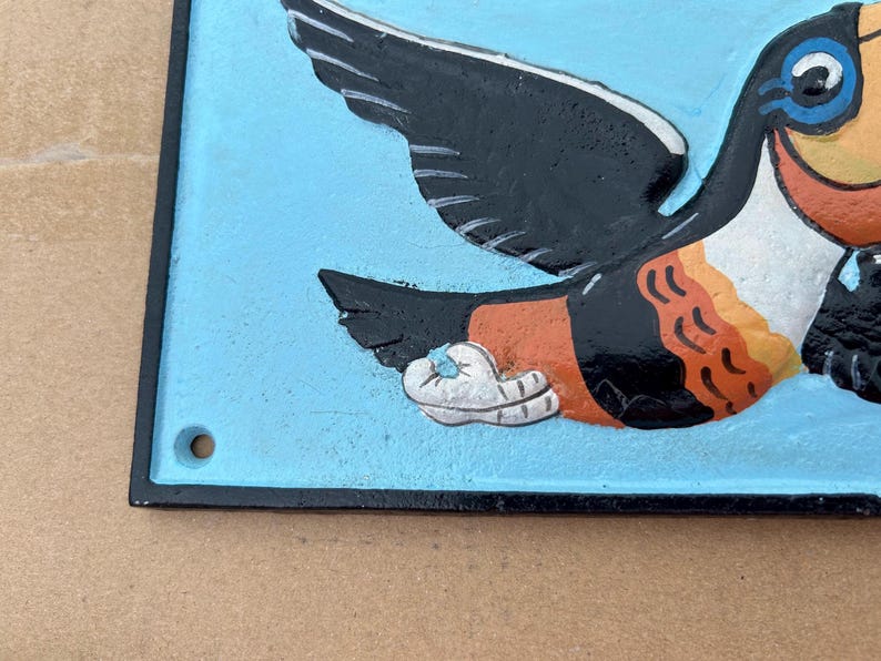 May include: A rectangular metal wall plaque with a light blue background featuring a relief illustration of a toucan. The bird has black wings, a white chest, and orange accents. A small circular hole is visible in the bottom left corner.