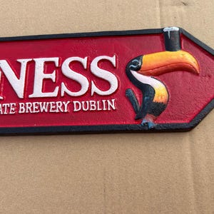 Op de afbeelding: Een rood en zwart pijl-vormig bord van gegoten metaal met de witte reli&euml;ftekst GUINNESS en ST. JAMES'S GATE BREWERY DUBLIN. Aan de rechterkant staat een kleurrijke toekan afgebeeld die een glas donker bier op zijn snavel in evenwicht houdt.