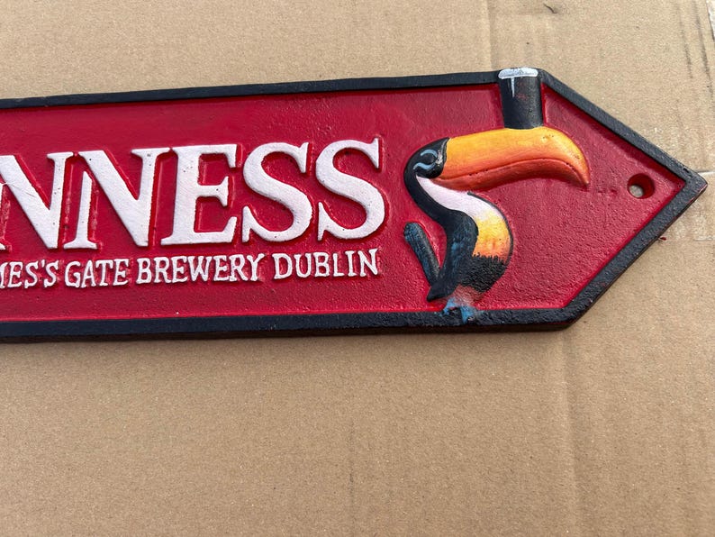 Op de afbeelding: Een rood en zwart pijl-vormig bord van gegoten metaal met de witte reli&euml;ftekst GUINNESS en ST. JAMES'S GATE BREWERY DUBLIN. Aan de rechterkant staat een kleurrijke toekan afgebeeld die een glas donker bier op zijn snavel in evenwicht houdt.