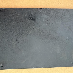 May include: A rectangular black metal plaque with a textured, matte finish. The plate features four small, circular mounting holes positioned near each of the four corners, allowing for secure attachment to a flat surface or wall structure.